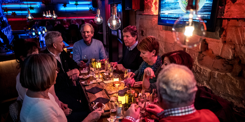 8 mensen zitten aan een lange houten tafel met hapjes en drankjes