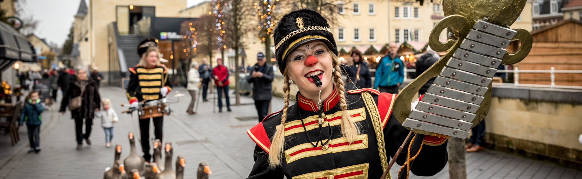 Vrouw en man in zwart rood fanfarepakje en rode neus met daartussen paraderende ganzen