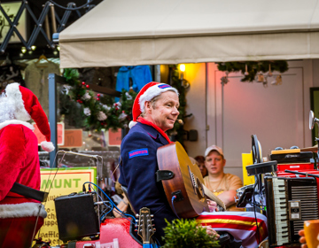 Man met kerstmuts en gitaar te midden van instrumenten voor terras. Kerstman loopt uit beeld